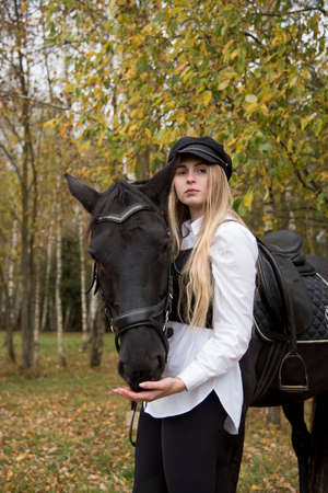 Portrait of a teenage girl with long blonde hair and a black cap, hugging a Chestnut horse against the background of an autumn forest.の写真素材