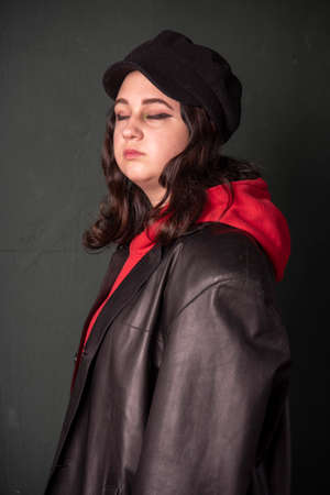 A girl in a black hat , wearing a red hoodie and a black leather jacket on the background of a dark wall. Photo shoot in the studio. Youth trend.の写真素材
