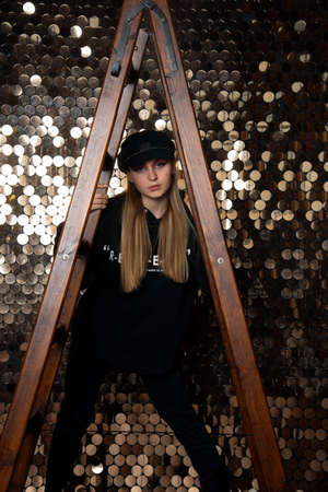 Womens photo shoot. A teenage girl in black clothes holds on to a stepladder against a golden wall.の写真素材