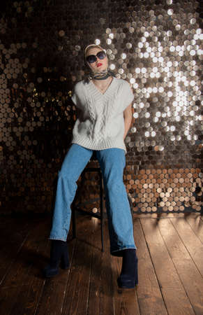 A girl in a white knitted vest, dark glasses and a silk scarf against the background of a golden wall. Fashion trends, photo shoot in the studio.の写真素材