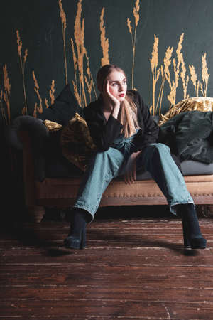 A girl in jeans and a black jacket sits on a vintage sofa against a dark wall. Photo shoot in a photo studio.の写真素材