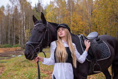 A slender girl with blonde hair stands next to a horse on the background of an autumn landscape.の写真素材