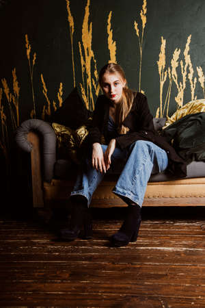 A girl in jeans and a black jacket sits on a vintage sofa against a dark wall. Photo shoot in a photo studio.の写真素材