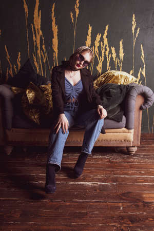A girl in jeans and a black jacket sits on a vintage sofa against a dark wall. Photo shoot in a photo studio.の写真素材