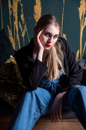 A girl in jeans and a black jacket sits on a vintage sofa against a dark wall. Photo shoot in a photo studio.の写真素材