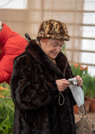 Russia, Moscow, February 2021.An elderly woman in a fur hat and fur coat stands in a flower greenhouse with a medical mask and a camera in her hands.のeditorial素材