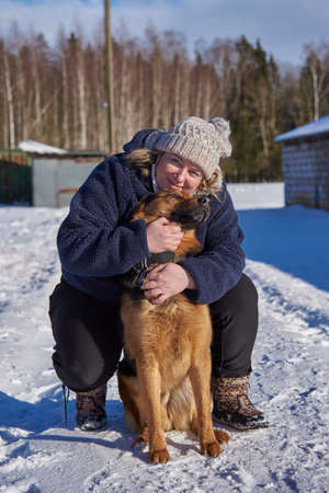 A plump middle-aged blonde woman in a knitted hat hugs a mongrel sitting on white snow on a sunny winter day.の写真素材