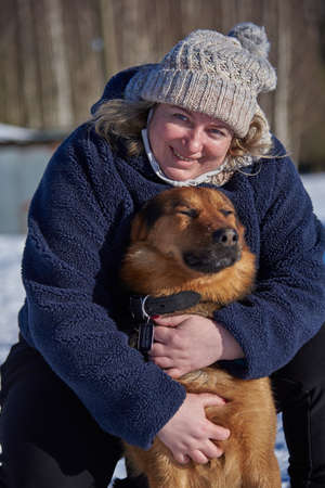 A plump middle-aged blonde woman in a knitted hat hugs a mongrel sitting on white snow on a sunny winter day.の写真素材