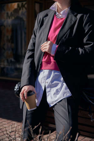 A woman in a black men's suit and white shirt with a disposable cup of coffee in her hands on a sunny day.の写真素材