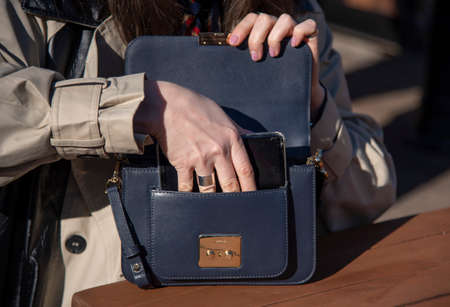 A woman in a light raincoat puts her phone in her purse, sitting in an open cafe on a sunny day.