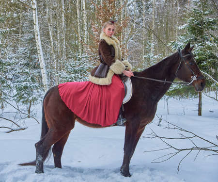 A girl in a beautiful old-fashioned burgundy dress on a horse in a winter forest.の写真素材