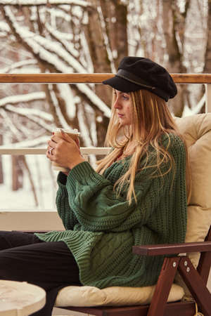 A blonde girl is sitting at a table on the veranda against the background of a winter snow-covered park.の写真素材