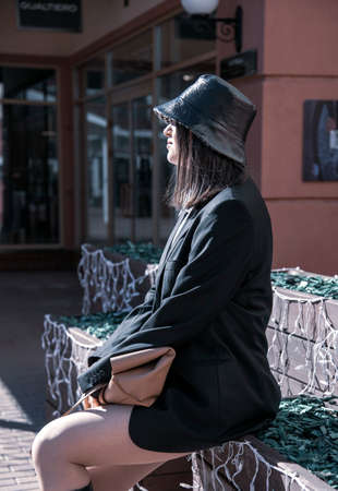 A woman in a black hat, a black man's jacket, and a very short skirt sits on a bench on a sunny spring day.の写真素材