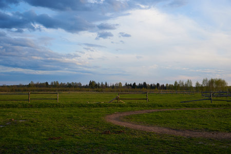 A green field with a wooden fence for horses against the background of a sunset cloudy sky. Spring landscape.の写真素材