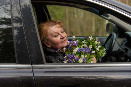 An elderly woman of 65 years sits in a black car with a bouquet of forest spring flowers.の写真素材