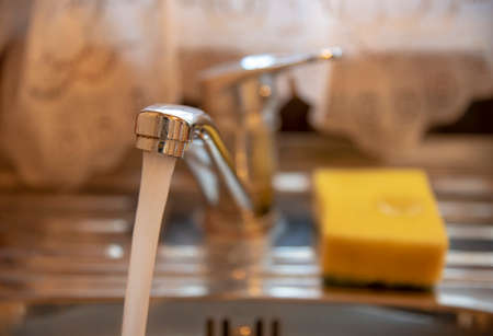 Kitchen faucet with running water. On the sink is a yellow used sponge.の写真素材