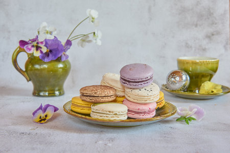 Gentle still life. Several colorful macaroons on a ceramic saucer, a cup of tea and a jug of flowers in the background.の写真素材