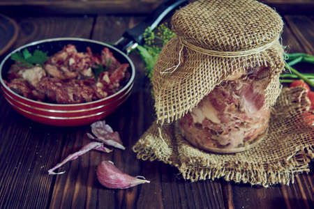 A jar of stewed meat and a frying pan, various vegetables and greens on a wooden countertop.の写真素材
