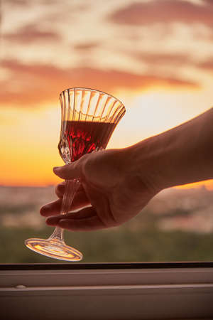 A man's hand with a glass of red wine on the background of a bright sunset.の写真素材