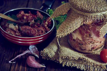 A jar of stewed meat and a frying pan, various vegetables and greens on a wooden countertop.
