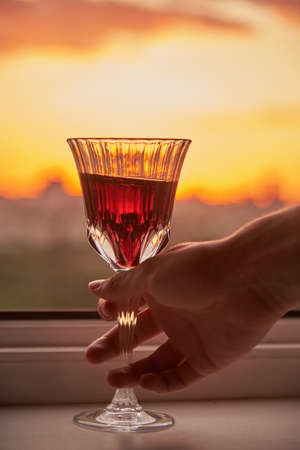 A man's hand with a glass of red wine on the background of a bright sunset.の写真素材