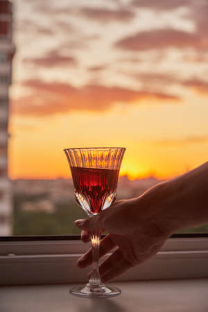 A man's hand with a glass of red wine on the background of a bright sunset.の写真素材
