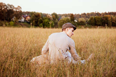 An 18-year-old boy in a cap is sitting with a dog on a field in the thick grass on a summer evening.の写真素材