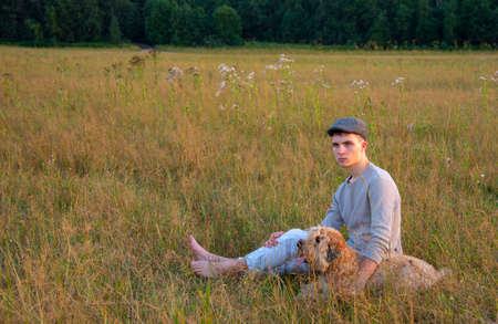 An 18-year-old boy in a cap is sitting with a dog on a field in the thick grass on a summer evening.の写真素材