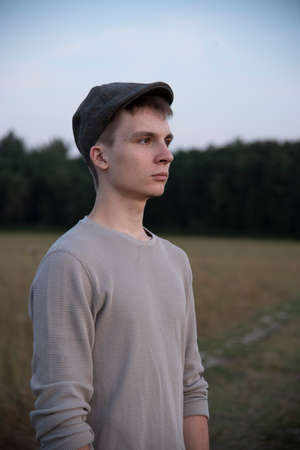 Portrait of an 18-year-old boy in a cap, standing on a dirt road in the middle of a field.の写真素材