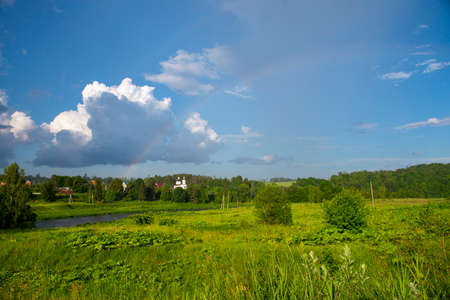 Bright sunny summer landscape with a rainbow. An elegant church in the middle of green trees. The river flows among the fields.の写真素材