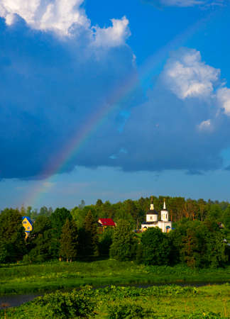 Bright sunny summer landscape with a rainbow. An elegant church in the middle of green trees. The river flows among the fields.の写真素材