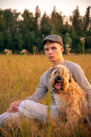 An 18-year-old boy in a cap is sitting with a dog on a field in the thick grass on a summer evening.の写真素材
