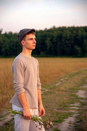 Portrait of an 18-year-old boy in a cap, standing on a dirt road in the middle of a field.の写真素材