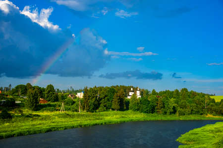 Bright sunny summer landscape with a rainbow. An elegant church in the middle of green trees. The river flows among the fields.の写真素材
