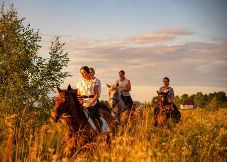 Russia, Ruza, August 2021. Girls in white shirts are riding horses on a blooming meadow in the rays of the setting sun.のeditorial素材