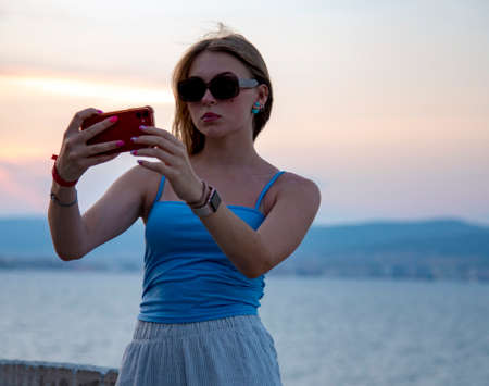 A blonde girl in dark glasses takes pictures of herself on her phone against the background of the sea and the sunset.の写真素材