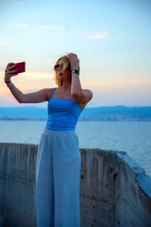 A blonde girl in dark glasses takes pictures of herself on her phone against the background of the sea and the sunset.の写真素材