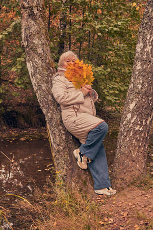 An elderly gray-haired woman in a light jacket is walking in the park with a bouquet of leaves.の写真素材