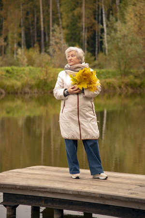 An elderly woman with a bouquet of yellow maple leaves, dressed in a light jacket, stands on a wooden bridge over a pond in an autumn park.の写真素材