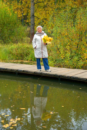 An elderly woman with a bouquet of yellow maple leaves, dressed in a light jacket, walks on a wooden deck over a pond in an autumn park.の写真素材