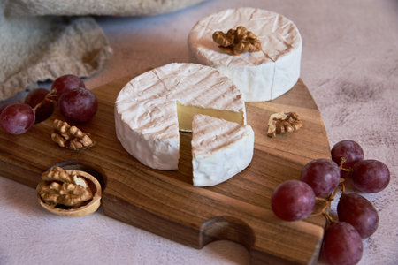 Cheese with white mold, grapes and walnuts on a wooden cutting board on a light countertop.の写真素材
