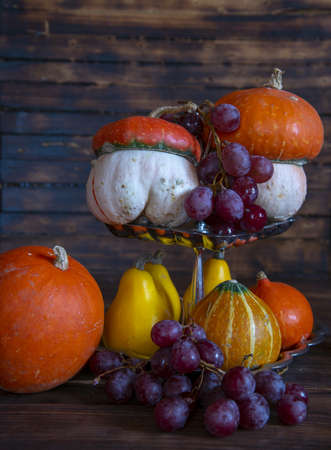 Pumpkins of different shapes, colors and sizes and delicious grapes on a silver stand on a wooden background. Autumn still life.の写真素材