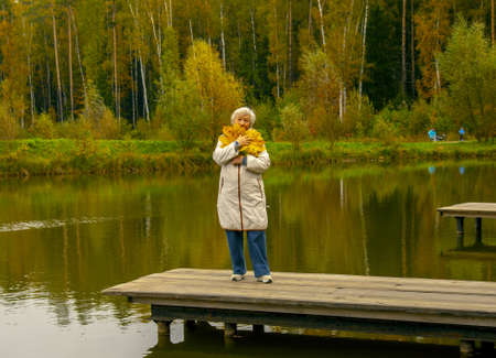 An elderly woman with a bouquet of yellow maple leaves, dressed in a light jacket, stands on a wooden bridge over a pond in an autumn park.の写真素材