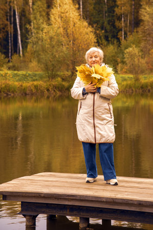 An elderly woman with a bouquet of yellow maple leaves, dressed in a light jacket, stands on a wooden bridge over a pond in an autumn park.の写真素材
