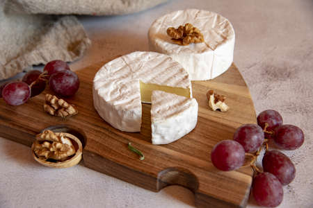 Cheese with white mold, grapes and walnuts on a wooden cutting board on a light countertop.の写真素材