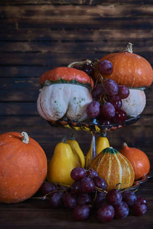 Pumpkins of different shapes, colors and sizes and delicious grapes on a silver stand on a wooden background. Autumn still life.の写真素材
