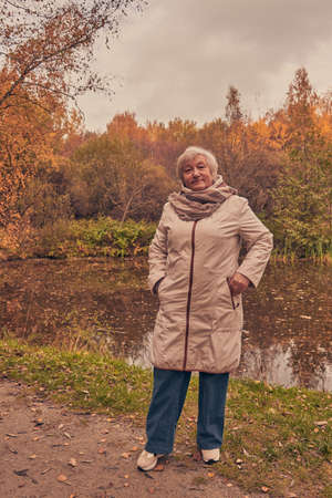 An elderly elegantly dressed woman stands on the shore of a pond on a cloudy autumn day.の写真素材