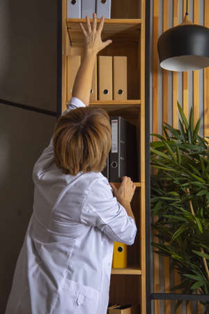 Russia, Moscow, December 2021. A female doctor pulls a cardboard folder from the shelf.の写真素材