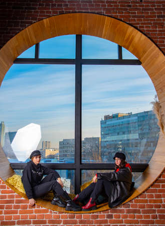 Two teenage girls are sitting on a wide windowsill by a large round window. Outside the window is a residential urban area.の写真素材