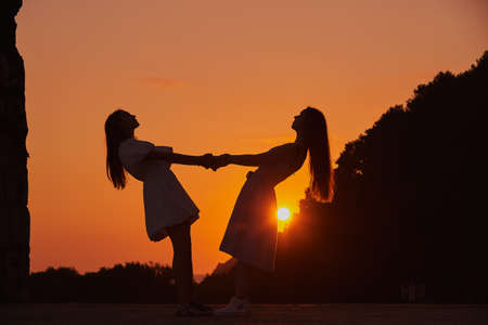 Silhouettes of two girls. holding hands against the background of the orange sky and the setting sun.の写真素材